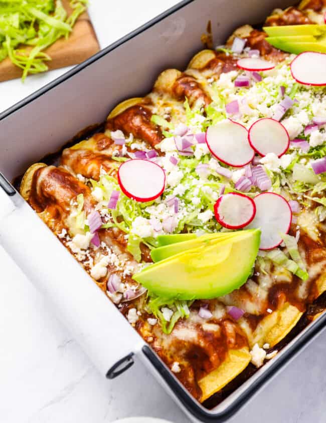 chicken enchiladas in a rectangular baking pan topped with lettuce, radish, onion, and avocado.