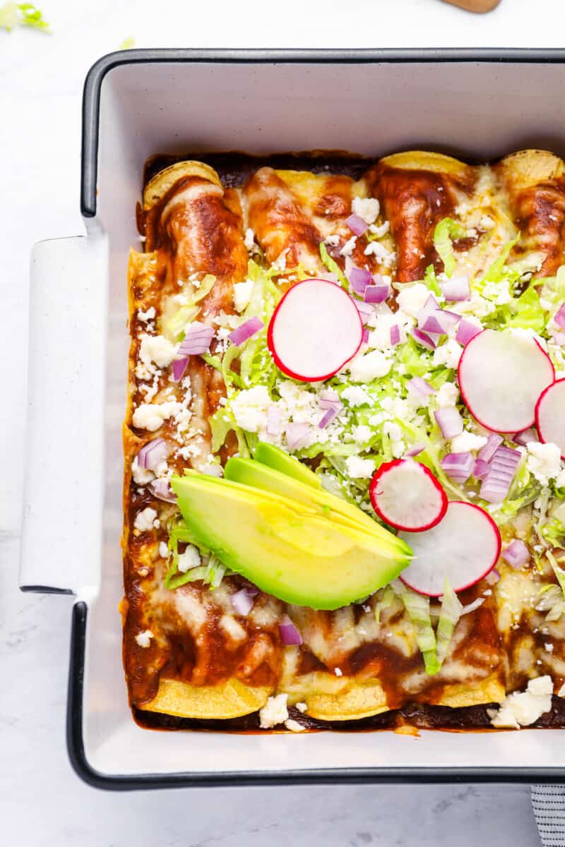 partial overhead view of chicken enchiladas topped with lettuce, radish, onion, and avocado.
