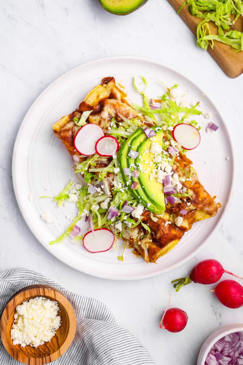 overhead view of 2 chicken enchiladas on a white plate with lettuce, radish, onion, and avocado.