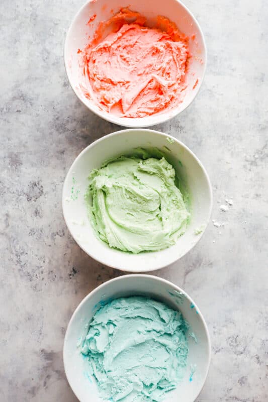 overhead view of cream cheese mint dough in 3 white bowls colored red, green, and blue.