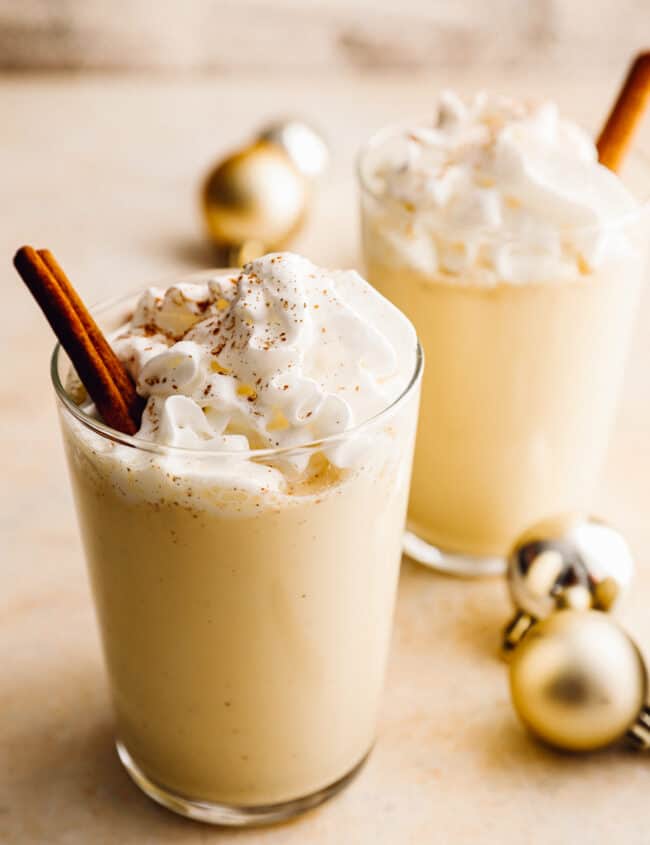 2 glasses of crockpot eggnog with whipped cream and cinnamon sticks.