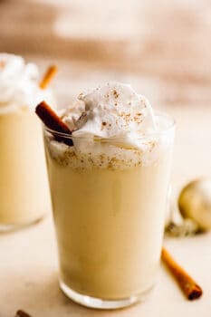 2 glasses of crockpot eggnog with whipped cream and cinnamon sticks.