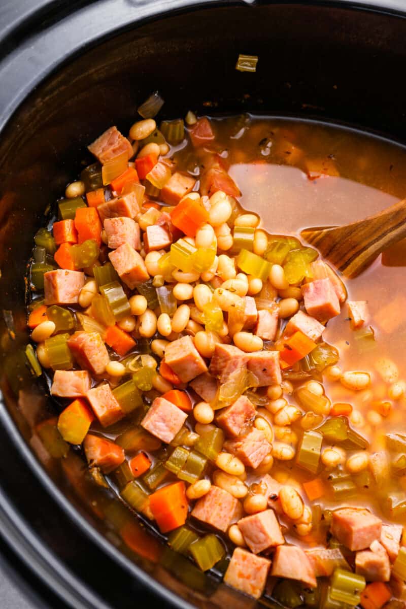 close up of crockpot ham and bean soup in a crockpot with a wooden spoon.