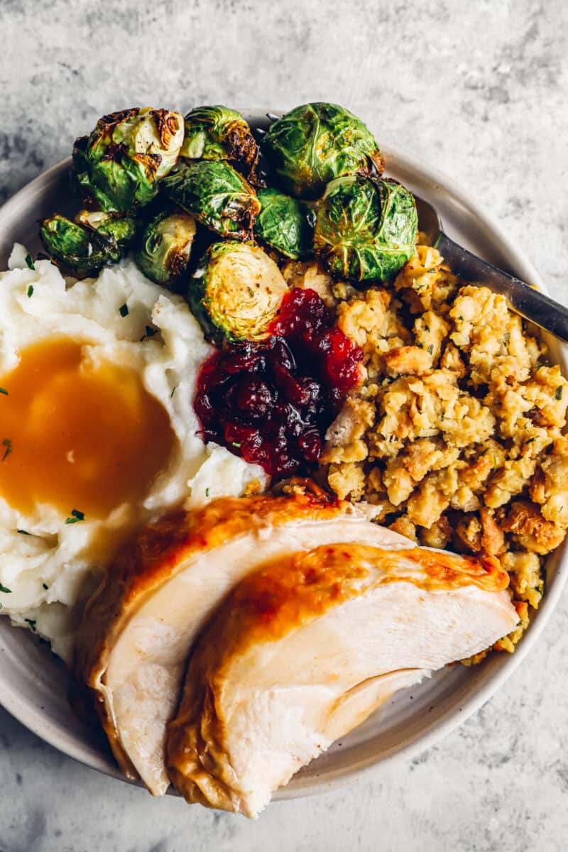 deep fried turkey slices on a white plate with stuffing, mashed potatoes, gravy, brussels sprouts, and cranberry sauce.