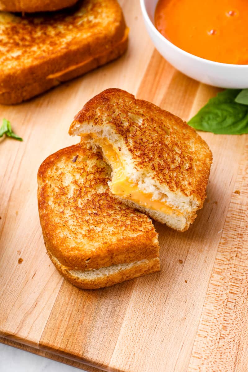 a bitten half of a grilled cheese stacked on the other half on a wooden cutting board.