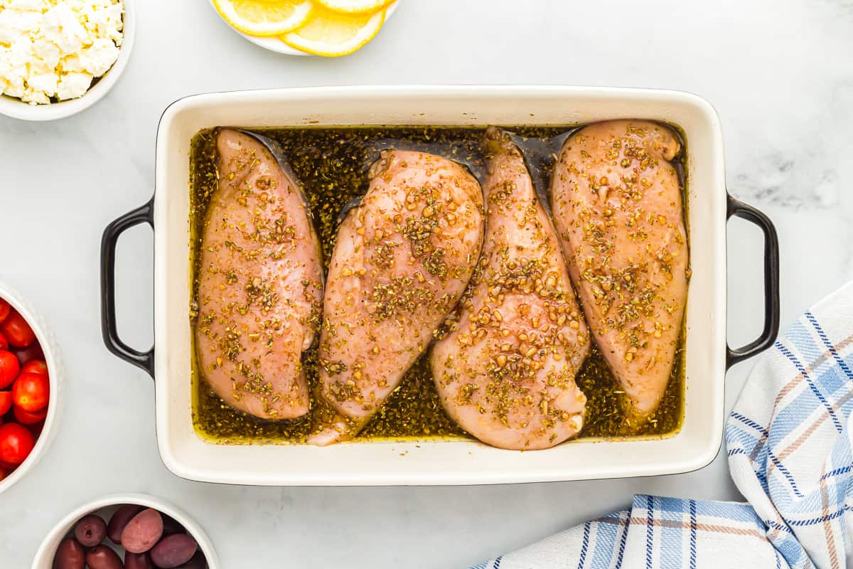 4 raw chicken breasts in a rectangular baking dish with greek marinade poured over them.