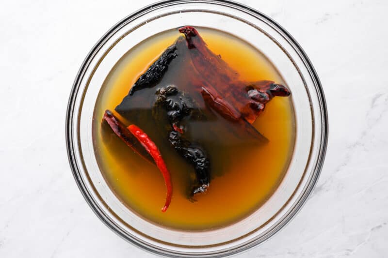 dried chilis soaking in chicken broth in a glass bowl.