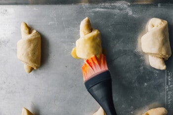 a pastry brush brushing egg wash over unbaked croissants on a baking sheet.