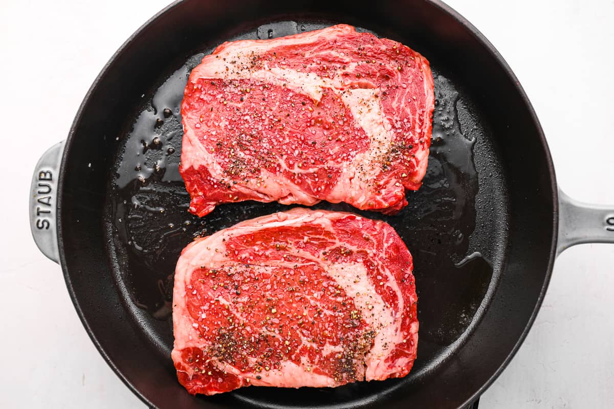 2 seasoned ribeye steaks searing in a cast iron pan.