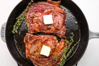 2 seared ribeye steaks topped with butter and thyme in a cast iron skillet.