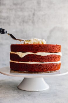 a spatula spreading cream cheese frosting over 3 stacked layers of red velvet cake on a white cake stand.