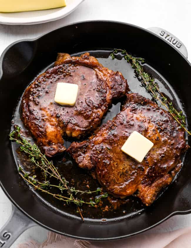 overhead view of oven baked steaks in a cast iron pan topped with a pat of butter and thyme.