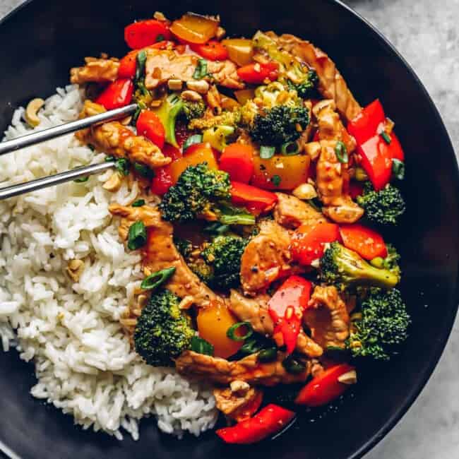 metal chopsticks grabbing a piece of pork from pork stir fry with white rice in a black bowl.