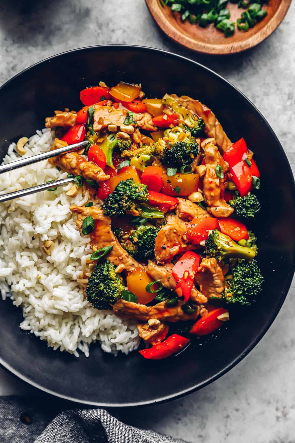 metal chopsticks grabbing a piece of pork from pork stir fry with white rice in a black bowl.