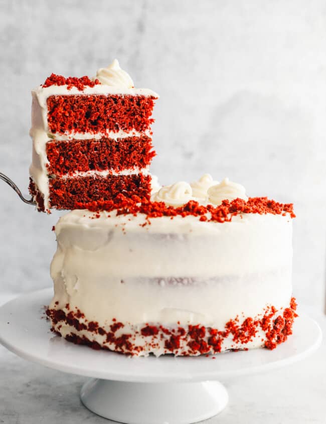 a cake server lifting a slice of red velvet cake from a cake on a white cake stand.