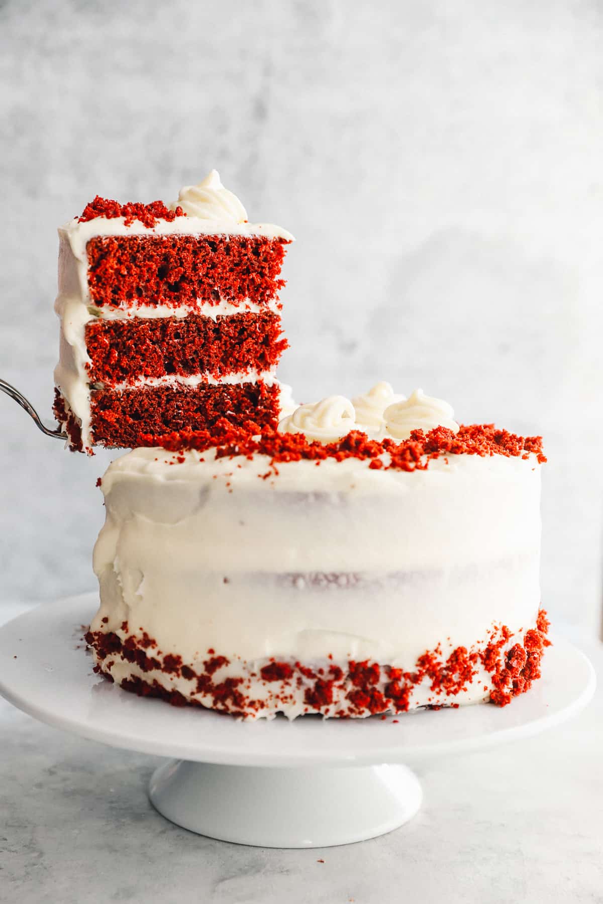 a cake server lifting a slice of red velvet cake from a cake on a white cake stand.