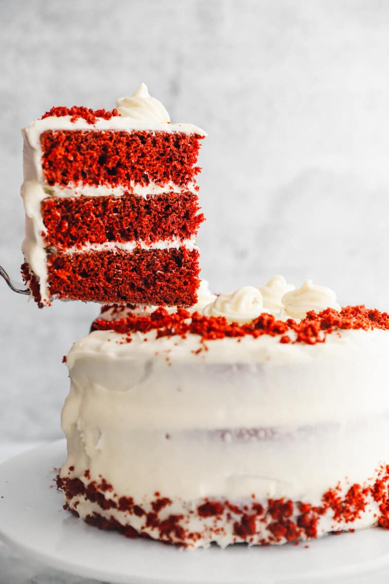 a cake server lifting a slice of red velvet cake from a cake on a white cake stand.