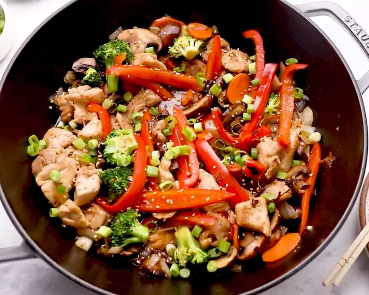 Finished chicken stir fry in a skillet with broccoli, peppers, mushrooms, green onions, and sesame seeds