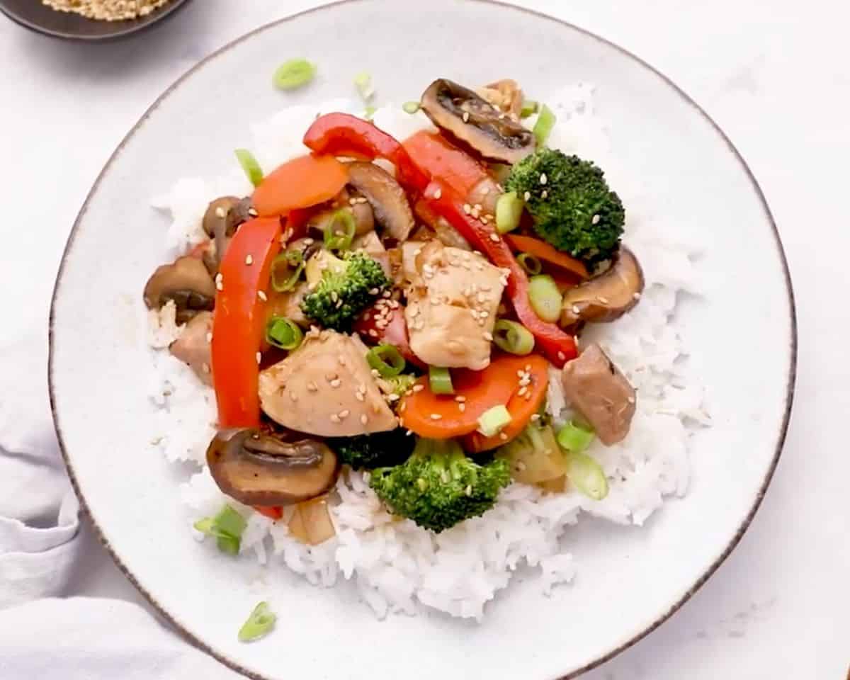 Chicken stir fry served over white rice in a bowl, garnished with sesame seeds and green onions