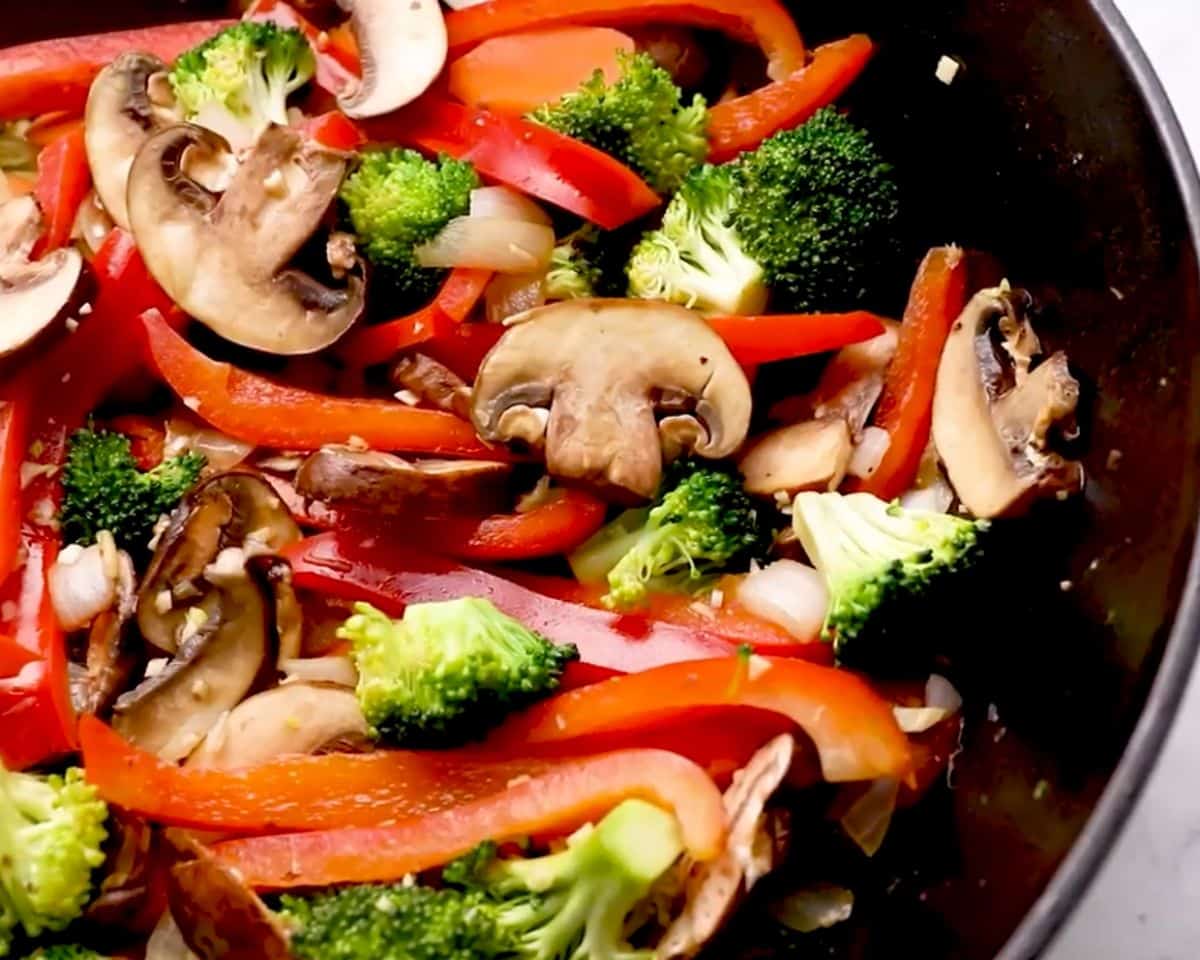 Fresh broccoli, red bell peppers, mushrooms, carrots, and onions sautéing in a skillet for homemade chicken stir fry