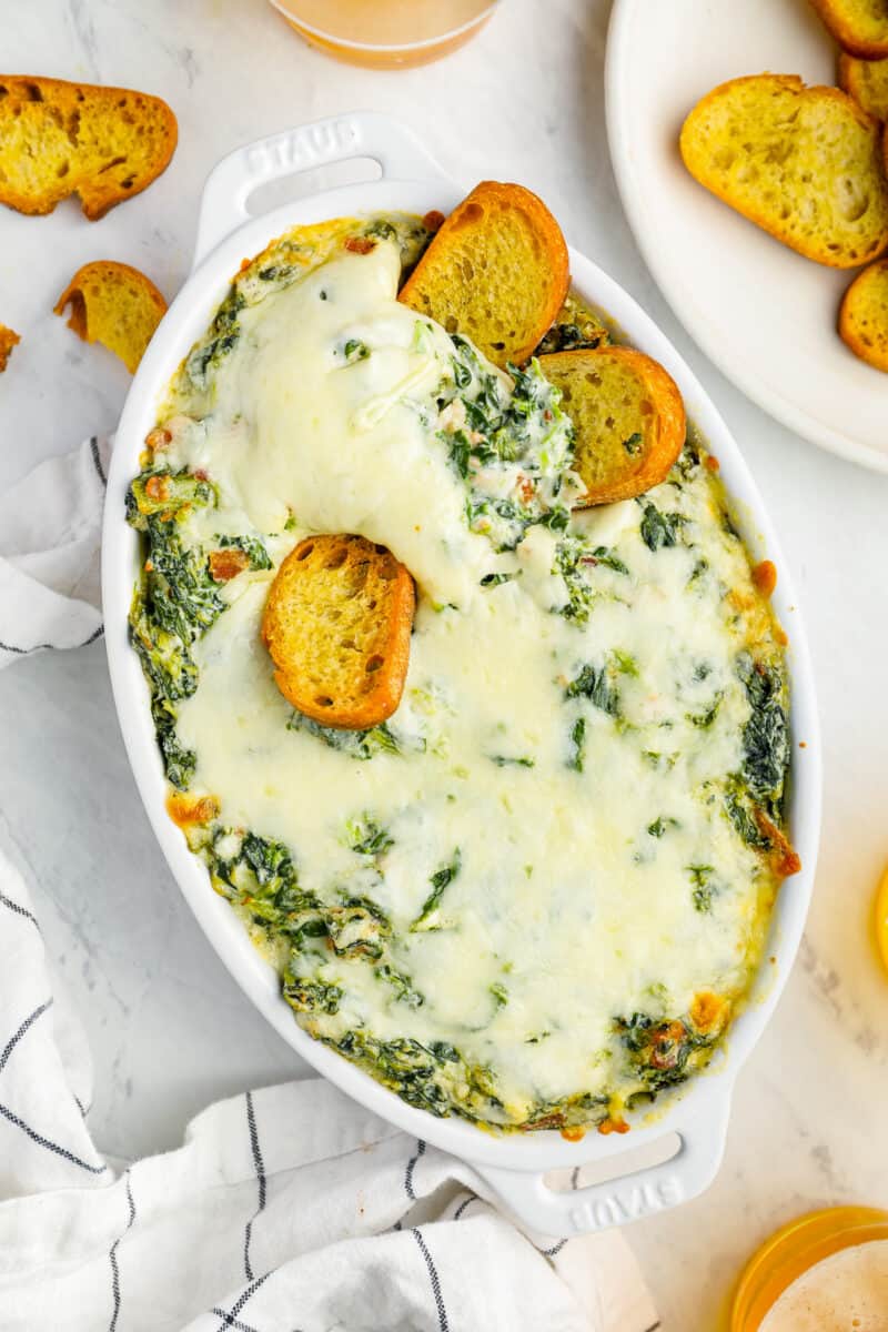 overhead view of bacon spinach dip in an oval baking dish with toasted bread pieces stuck in it.