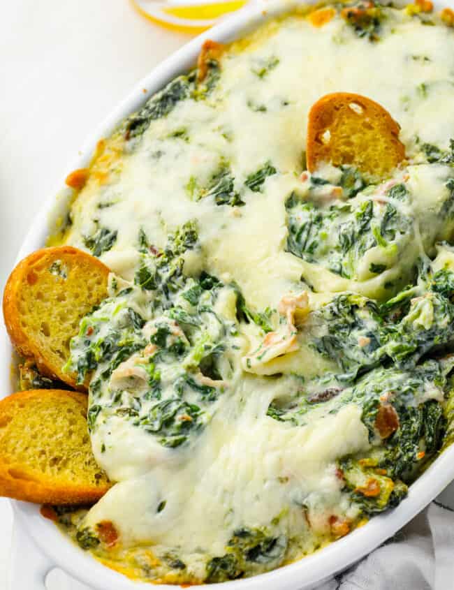 bacon spinach dip in an oval baking dish with toasted bread pieces stuck in.