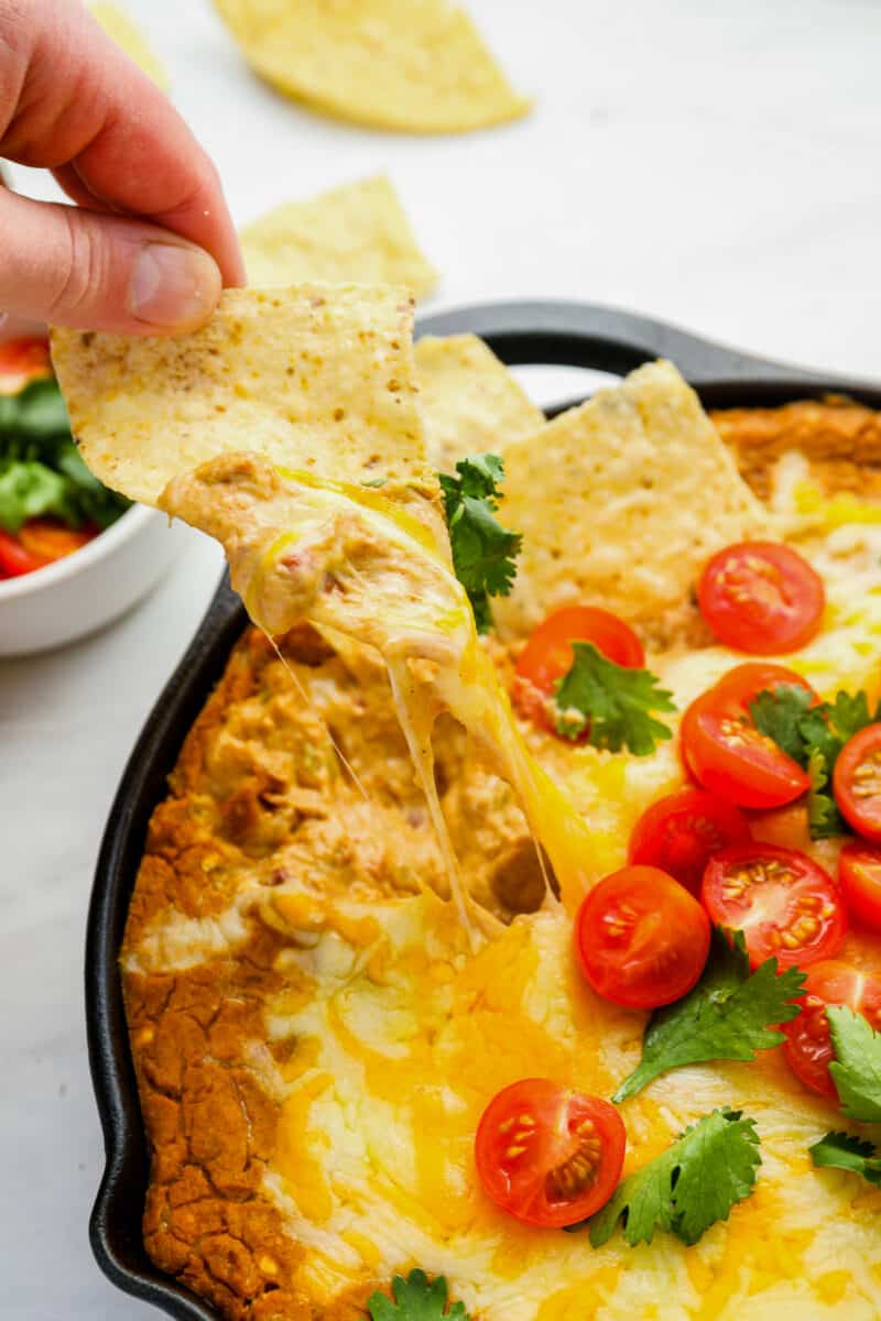 a hand using a chip to scoop cheesy bean dip from a cast iron pan.
