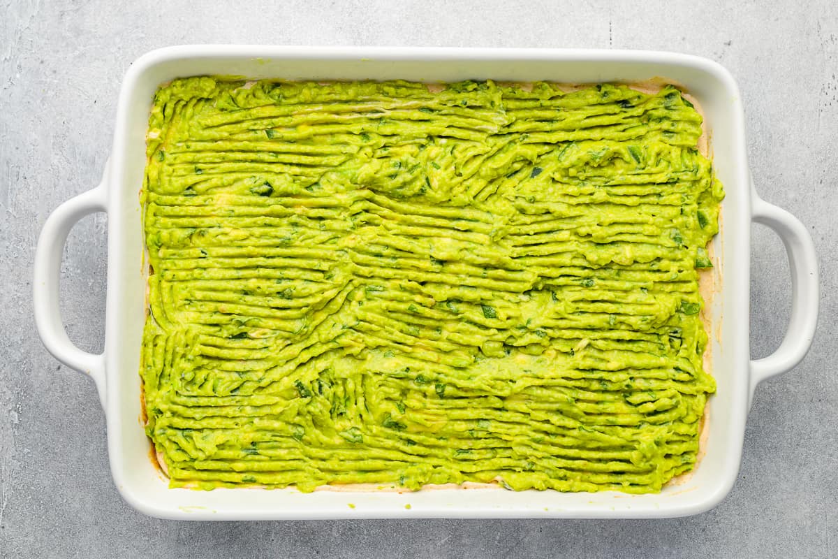 Guacamole spread over sour cream layer in a rectangular baking dish.