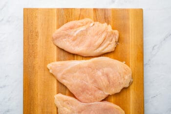 3 thin raw chicken breasts on a wooden cutting board.
