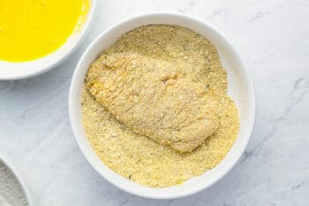 a raw chicken breast being dredged in seasoned breadcrumbs in a white bowl.