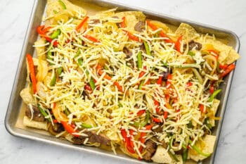 shredded cheese sprinkled over philly cheesesteak nachos on a baking sheet.