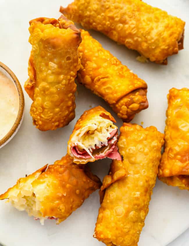 6 reuben egg rolls on a white plate, one broken in half to expose the filling.