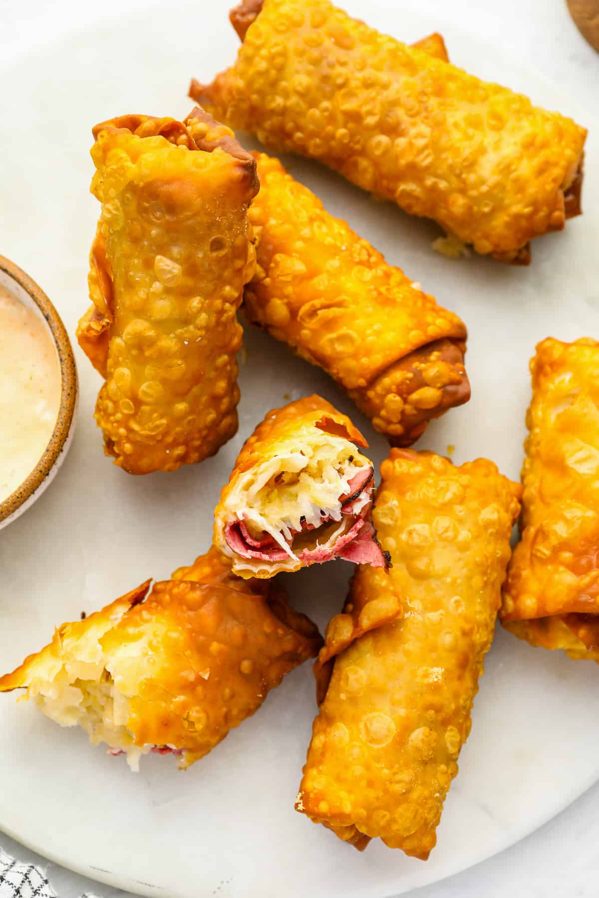 6 reuben egg rolls on a white plate, one broken in half to expose the filling.