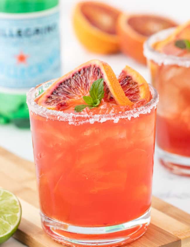 close up three-quarters view of a blood orange margarita on a wooden cutting board garnished with a blood orange slice and mint leaves.