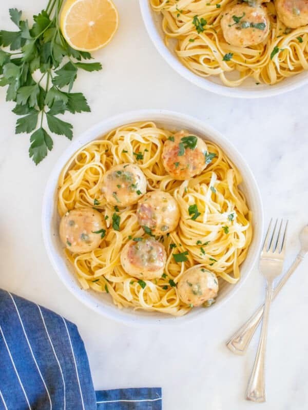 overhead view of 6 chicken piccata meatballs on a bed of fettuccini in a white bowl.