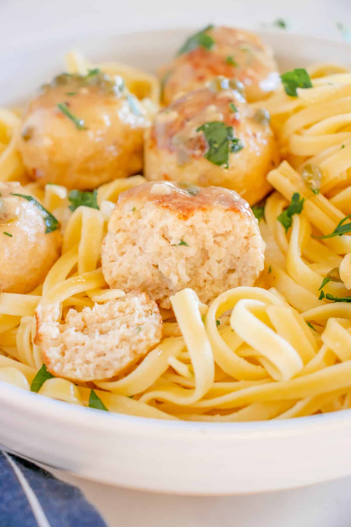 three-quarters view of a cut chicken meatball on a bed of fettuccini pasta with more whole meatballs in a white bowl.