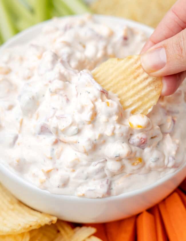 three-quarters view of a hand dipping a wavy potato chip into fiesta ranch dip in a white bowl surrounded by wavy potato chips, carrot sticks, tortilla chips, and celery sticks.