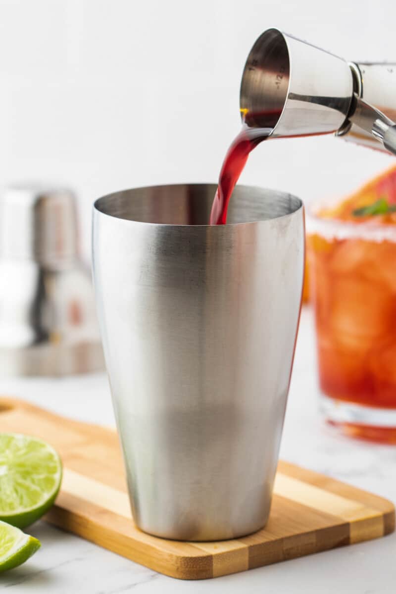 Blood orange juice being poured into a stainless steel shaker on a wooden cutting board.