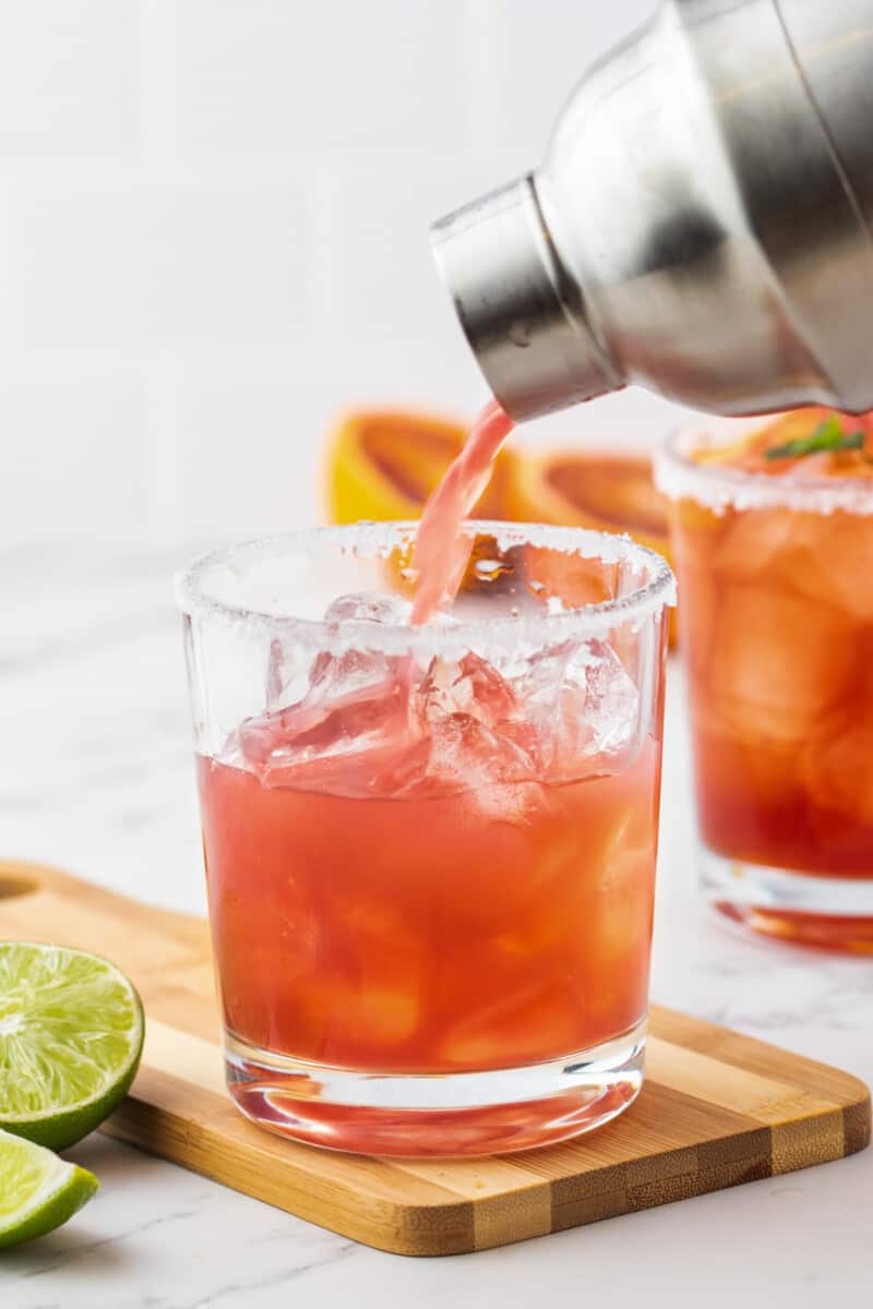 A blood orange margarita being poured from a stainless cocktail shaker into a rimmed short rocks glass with ice on a wooden cutting board.
