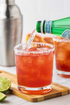 seltzer poured over a blood orange margarita in a rimmed short rocks glass on a wooden cutting board.