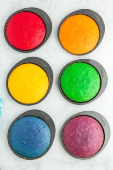 overhead view of baked rainbow cake layers in round cake pans, colored red, orange, yellow, green, blue, and purple.