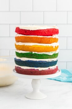 side view of stacked rainbow layers of cake on a white cake stand.