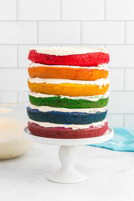 side view of stacked rainbow layers of cake on a white cake stand.