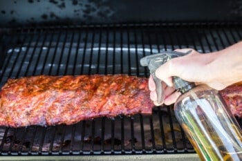 a hand spraying partially smoked ribs on a grill with apple juice.