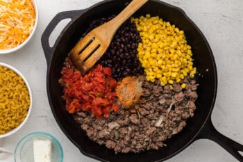 corn, beans, tomatoes, and seasonings added to ground beef and onion in a cast iron pan with a wooden spoon.