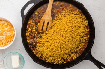 pasta added to taco mac and cheese in a cast iron pan with a wooden spoon.