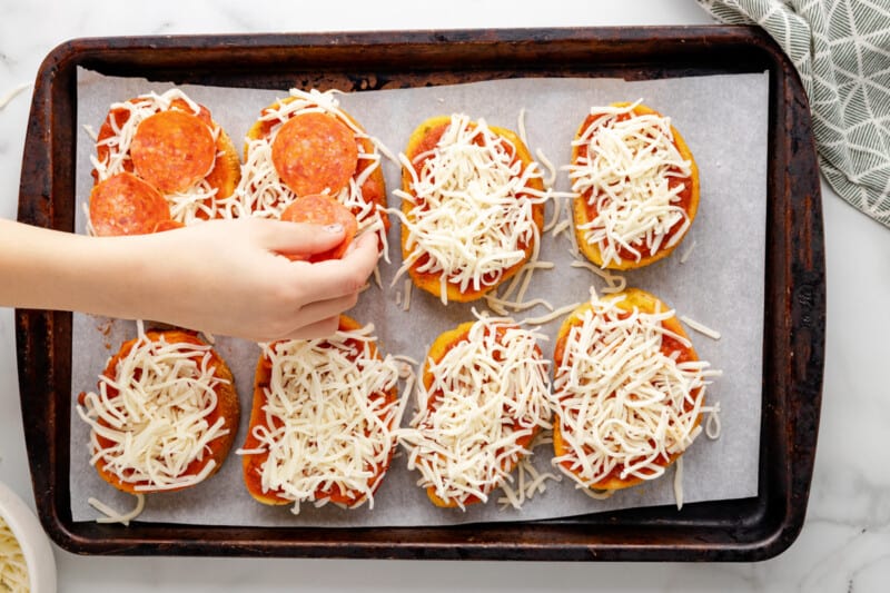a hand adding slices of pepperoni to texas toast pizza on a baking sheet.