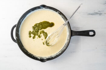 pesto added to cheese sauce in a cast iron pan with a whisk.
