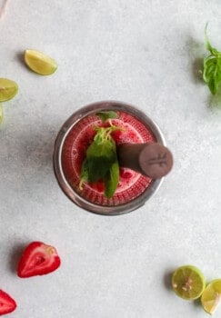 strawberries, basil, and lime juice being muddled in a glass cocktail shaker.