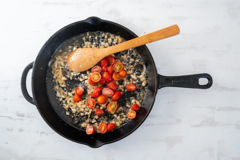 tomatoes added to garlic and onion in a cast iron pan with a wooden spoon.